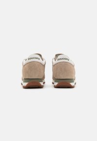 Beige semskede sneakers med en hvid hæl-tab med logo, grønne detaljer og en struktureret gummisål. Klassisk sporty design.