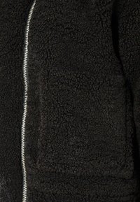 Čierna flísová bunda s textúrovaným povrchom, so strieborným zipsom na prednej strane a diskrétnou náprsníkovou vreckom na ľavej hrudi.