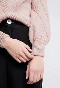 Lyserød strikket sweater med et struktureret diamantmønster, der har ribbede manchetter og en afslappet pasform, båret over sorte bukser med knapdetaljer.