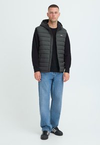 Tume roheline puffer vest koos kapuutsiga, lukuga ja küljetaskutega, musta sviitri peal, koos lahtiste siniste teksade ja mustade tossudega.