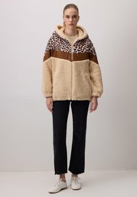 Chaqueta de felpa sintética color tostado con un diseño de estampado animal y paneles marrones, con capucha y bolsillos laterales. Combinada con pantalones negros y zapatos blancos.