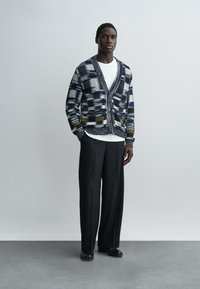 Mężczyzna stoi w wzorzystym kardiganie zapinanym na guziki, białym t-shircie, czarnych szerokich spodniach i czarnych butach na jednolitym tle.