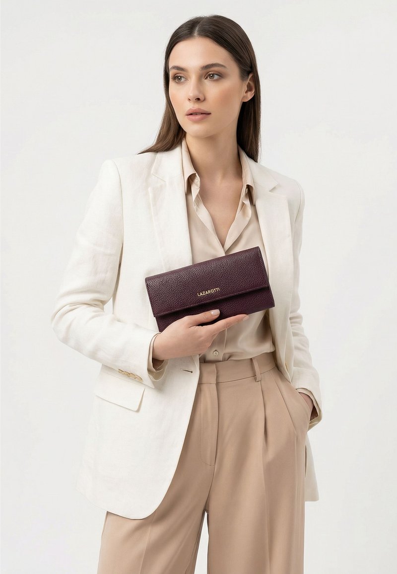 Femme portant un blazer blanc et un pantalon beige tenant une pochette en cuir marron foncé avec "LAZAROTTI" embossé en or.