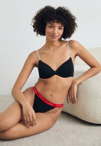 Svart lingerie-set bestående av en sömlös bh med tunna vita logoband och svarta höga trosor med ett rött logoband.