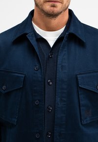Marineblaue Arbeitsjacke mit zwei Brusttaschen, Knopfverschluss und einem strukturierten Strick darunter. Glattes Material mit einem strukturierten Kragen.
