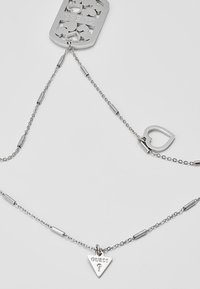 Collier chaîne en argent avec un pendentif triangulaire "GUESS?", un petit charme en forme de cœur ouvert et une étiquette décorative rectangulaire.