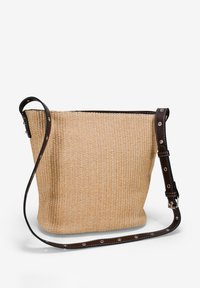 Bolso de bucket de paja trenzada en beige claro, que presenta una correa de cuero negro con acentos metálicos, superficie texturizada y un diseño de apertura superior.