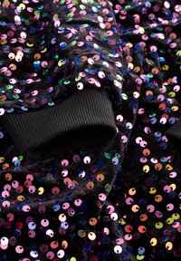 Tissu noir orné de sequins multicolores en formes circulaires, avec des poignets noirs côtelés. Les sequins reflètent la lumière, créant un effet scintillant.