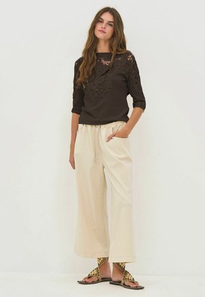 Femme portant un top en dentelle noire, un pantalon crème à jambes larges, des sandales dorées cloutées, debout avec une main dans la poche sur un fond uni.