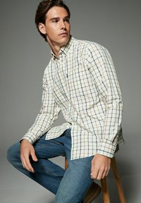 Chemise à manches longues à boutons blanche avec un motif à carreaux multicolore, dotée d'une poche poitrine et associée à un jean bleu.