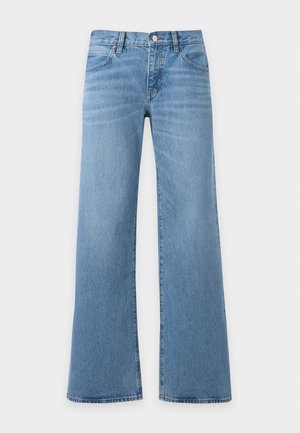 Femme portant un top marron à manches longues, un jean bleu à jambes larges, des sandales noires à talons, tenant une grande pochette en cuir marron.