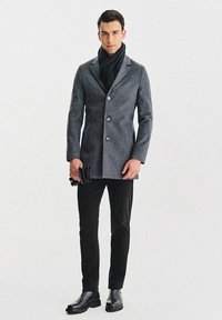 Szary wełniany płaszcz o dopasowanym kroju, jednorzędowy z guzikami i kołnierzem z klapą. Noszony z czarnymi spodniami i błyszczącymi czarnymi butami.