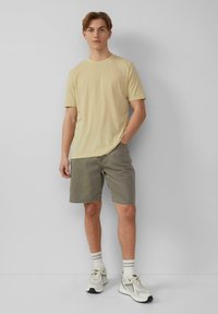 Jasnożółta bawełniana koszulka, luźny krój, okrągły dekolt; oliwkowe szorty; szaro-białe buty sportowe w paski z skarpetkami; prosty design.