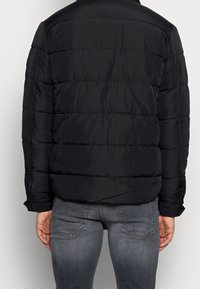 Svart vadderad jacka med quiltad yta, hög krage och långa ärmar. Synlig sömnad och knäppta ärmslut. Bärs med denimjeans.