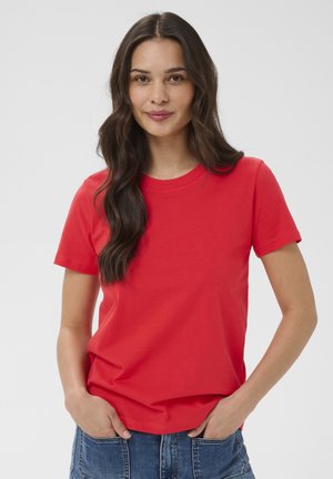 Kvinde med langt mørkt hår iført en simpel rød t-shirt med korte ærmer og blå jeans, stående med hænderne i lommerne.