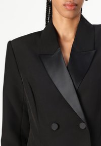 Musta bleiseri, jossa on satiininen huippuaplikaatio, kaksinappinen suljenta ja jämäkät olkapäät. Silkkinen kangas, jossa on matta pinta.