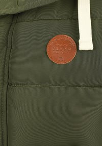 Olivgrön vadderad jacka med texturerat tyg, med en rund brun läderlapp märkt "BLEND" och en vit dragsko vid huvan.