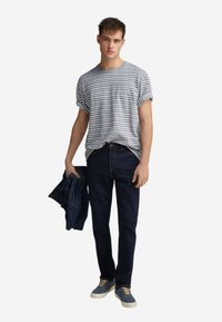 Pruhované šedé a bílé tričko s krátkým rukávem, tmavě modré džíny, modré boty se světlými šňůrkami; drží modrou džínovou bundu.