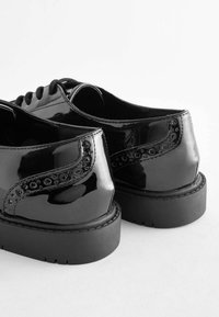 Chaussures brogue noires brillantes avec des semelles en caoutchouc épaisses, ornées de motifs perforés, présentées sur un fond clair.