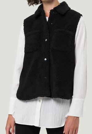 Black fleece button-up vest with two chest pockets worn over a long-sleeve white textured shirt and black pants.