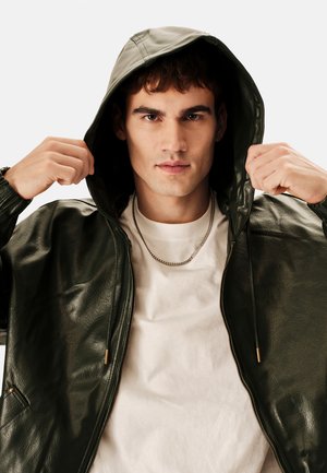 Giacca con cappuccio in ecopelle verde con zip frontale, polsini elastici e due tasche laterali, indossata sopra una camicia bianca con una collana a catena.