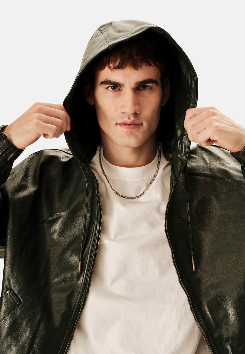 Chaqueta con capucha de cuero sintético verde, con cremallera frontal, puños elásticos y dos bolsillos laterales, combinada con una camisa blanca y un collar de cadena.