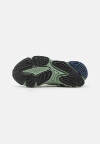 Podešev boty v zelené a černé gumě; obsahuje různé texturované vzory a modrý akcent na patě pro další designový prvek.