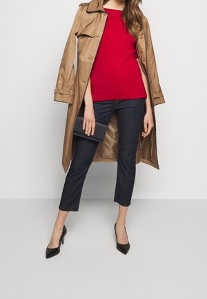 Kvinna i en beige trench coat över en röd topp och mörka avskurna jeans, som håller en svart kuvertväska och bär svarta högklackade skor.