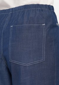 Pantaloni in denim blu scuro con vita elastica. Presentano una sola tasca posteriore con cuciture bianche e una superficie liscia.
