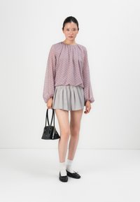 Young woman wearing a sheer patterned lavender blouse, gray pleated skirt, white socks, black flats, holding a small black handbag.