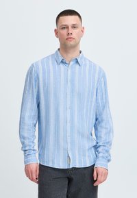 Chemise à manches longues à rayures bleu clair en tissu texturé avec col boutonné et manches retroussées, présentant des rayures verticales blanches.