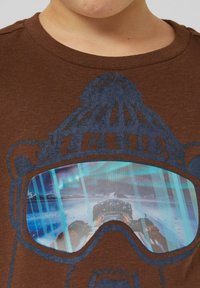 Braunes Baumwoll-T-Shirt mit einem Motiv aus blauen Skibrillen, die eine Schneelandschaft widerspiegeln, und einem darüber liegenden Hut-Design in dunklerem Blau.