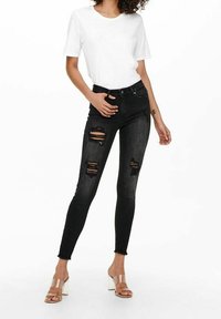 Pantalones vaqueros negros ajustados con desgastes en las rodillas, combinados con una camiseta blanca. La modelo lleva sandalias de tacón bloque transparente.