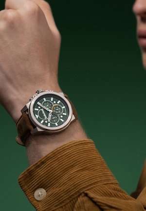 Montre chronographe avec un boîtier rond en argent, un cadran vert et un bracelet en cuir marron. Ses caractéristiques incluent plusieurs sous-cadrans et des marqueurs d’heure blancs.