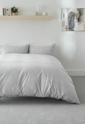 Bett mit hellgrauem Bettbezug und Kissen in einem minimalistischen Schlafzimmer mit einem Holzregal, das Bücher, Pflanzen und Dekor darüber hält, sowie abstrakter Kunst an der Wand.