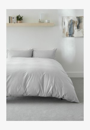 Bett mit hellgrauem Bettbezug und Kissen in einem minimalistischen Schlafzimmer mit einem Holzregal, das Bücher, Pflanzen und Dekor darüber hält, sowie abstrakter Kunst an der Wand.