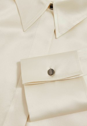 Cream satin shirt featuring a pointed collar, button closure, and a cuff with a black button accent. Smooth texture and structured design.