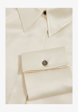 Chemise en satin crème avec col pointu, fermeture à boutons et poignet avec un accent de bouton noir. Texture lisse et design structuré.