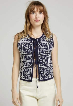 Gilet sans manches bleu marine avec broderie florale blanche en motifs carrés, présentant un col rond et une fermeture à nouer à l'avant.