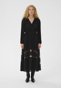 Vestido largo negro plisado con mangas largas y escote cruzado. Paneles transparentes crean un efecto de capas; tela texturizada en todo el vestido.