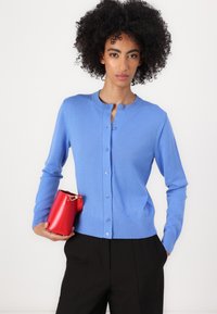 Cardigã azul claro com um decote redondo e fecho de botões; apresenta mangas longas e uma cintura ajustada. A modelo segura uma pequena bolsa vermelha.