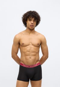 Man with curly hair standing shirtless wearing black Tommy Hilfiger boxer briefs with red waistband, hands behind back, neutral white background.