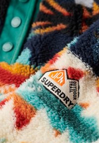 Gros plan sur une veste polaire colorée avec un patch logo Superdry et des boutons turquoise sur une patte en tissu bleu canard.