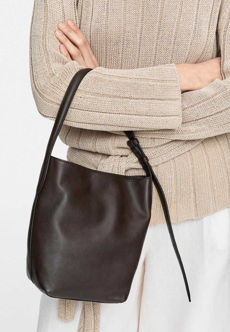 Personne en pull beige en tricot et pantalon blanc tenant un sac seau en cuir brun foncé avec une longue bandoulière sur les bras croisés.