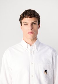 Jonge man met kort krullend haar en een wit overhemd met een klein zwart en gouden logo op de borstzak.
