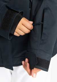 Marine waterdichte jas met elastische manchetten, klittenbandsluiting en zichtbare trekkoord. Het materiaal lijkt glad en heeft een zachte textuur.