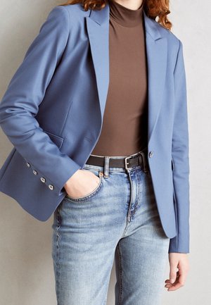 Persoon draagt een blauwe blazer over een bruine coltrui en lichtblauwe jeans met een zwarte riem, hand in de broekzak.
