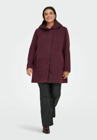 Burgundský vlněný kabát s velkou kapucí, předním zapínáním na knoflíky a postranními kapsami. Model má na sobě černé kalhoty a boty na podpatku.