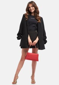 Czarna oversize marynarka, dopasowany czarny top, plisowana czarna spódniczka mini i złote sandały na ramiączkach. Niesie czerwoną skórzaną torebkę z łańcuszkiem.