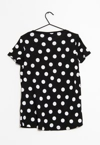 Camiseta negra de manga corta con cuello redondo y lunares blancos. La tela parece ligera y con una textura suave. Cuélga en un gancho negro.